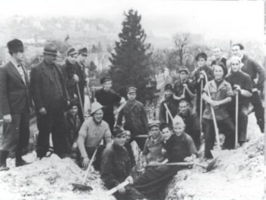 Ein Schwarz-Weiß-Foto eines Chors auf der Bühne. Der Chor besteht aus Männern und Frauen in festlicher Kleidung. Dahinter hängt ein Banner mit deutschem Text. Zuschauer sitzen davor und beobachten die Aufführung.